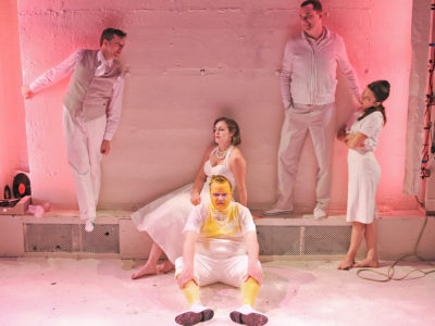 Two actors stand on a ledge laughing, two actors sit beneath them looking unhappy, one is covered in paint. A fifth actor stands and looks up at the laughing men.