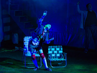 An actress dances in a blue spotlight, two lawn chairs are behind her.