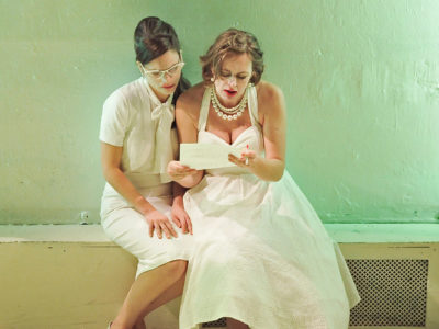 Two actresses wearing white 50s style costumes read a letter together on a bench.