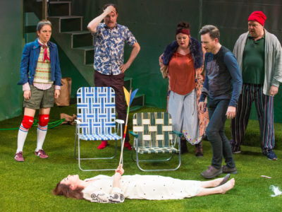 Four actors looking at (fifth actor) a woman in white dress lying in astro-turf with a toy arrow in her chest