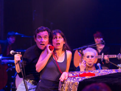 Two actors share a phone with confused expressions, behind them, a band plays.