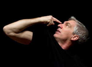 An actor in a scene: a man in black tshirt pointing at his own face with black background