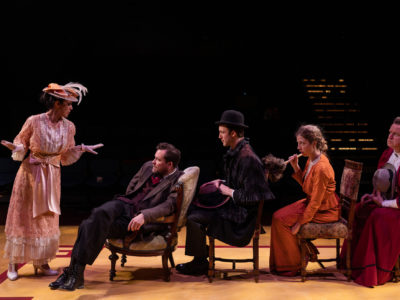 5 Actors on Stage, One standing the other sitting in single file row in chairs. Victorian Dress Costumes