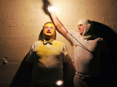 Two male actors, one covered in yellow paint, the other holding a lightbulb.