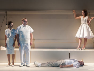 Five actors dressed in white in a surreal scene with one actor lying on the floor