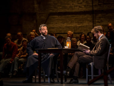 Two actors sitting at a table, one dressed as a priest, one reading a book, with audience in background