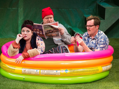 Three adult actors sitting in a rainbow colored inflatable baby pool, one biting her nails, one reading a newspaper, one playing cards