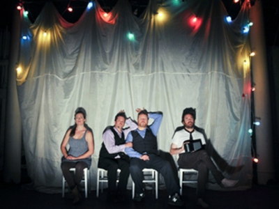 Four actors on a stage sitting in chairs with christmas lights above them