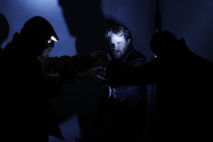 Four actors in dark setting hands all grasping a glowing indiscernible object that lights up the face of the central actor